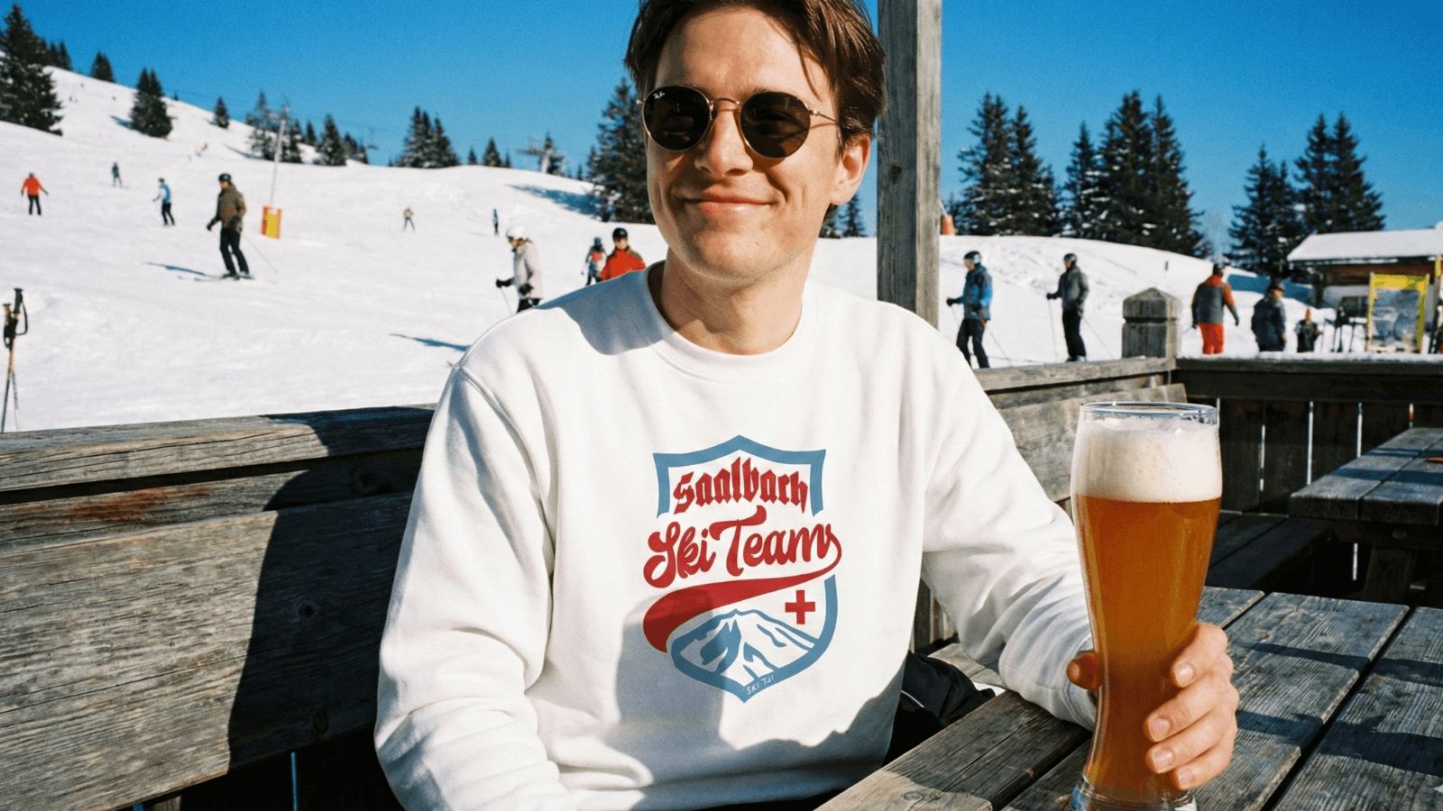 Person enjoying a beer while wearing a Saalbach Ski Team sweatshirt, with skiers in the background at a snowy mountain.