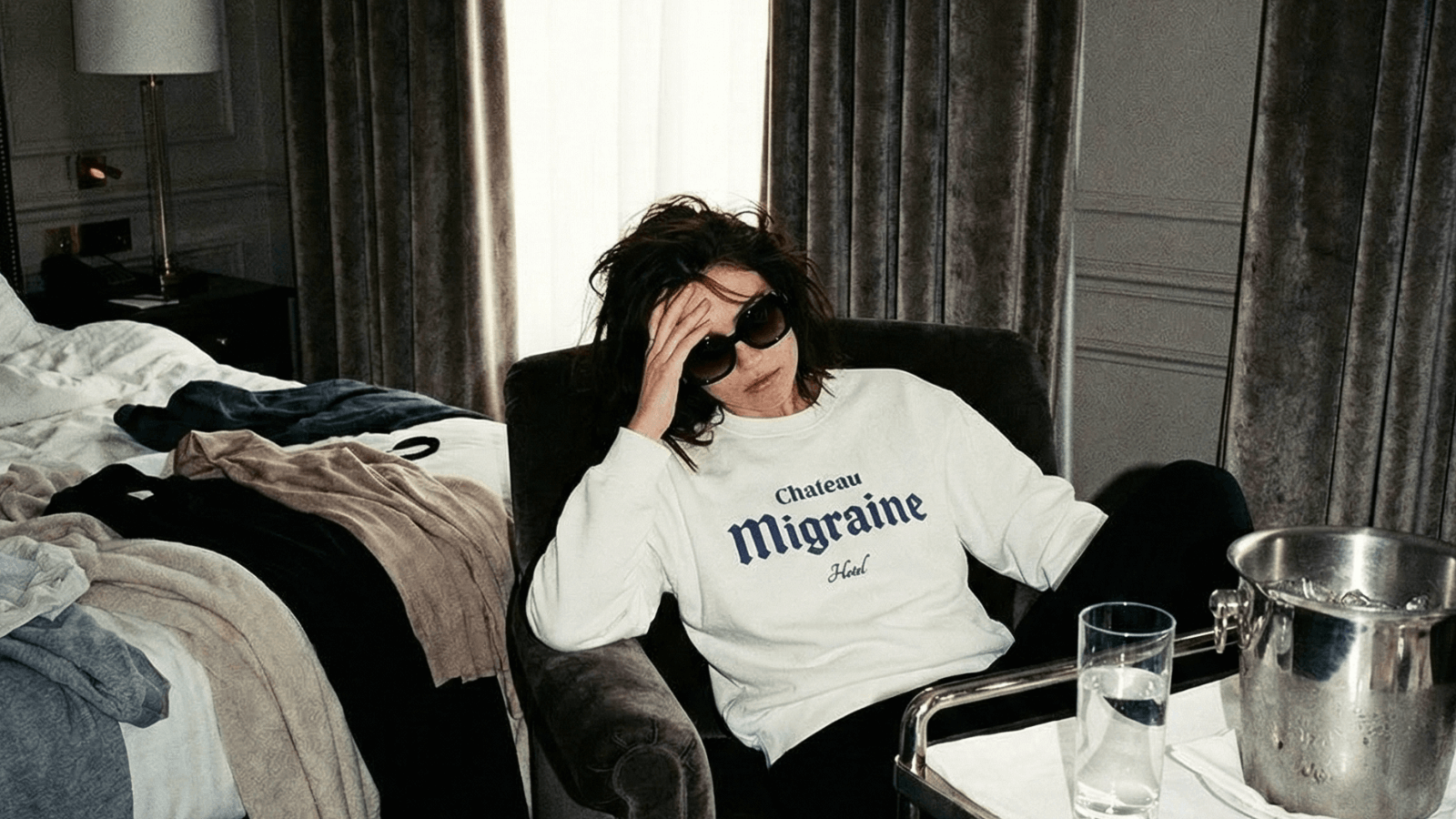 A person wearing a Chateau Migraine sweatshirt looking distressed in a hotel room, embodying the theme of CHATEAU MIGRAINE | FIVE STARS. ZERO MERCY!
