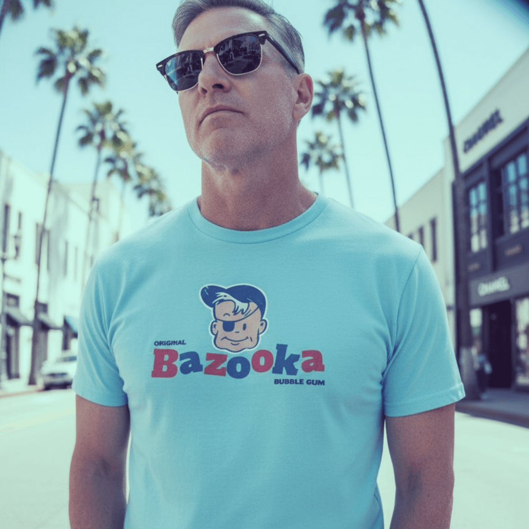 Man wearing a blue Bazooka Bubble Gum t-shirt, sunglasses, standing on a sunny street lined with palm trees.