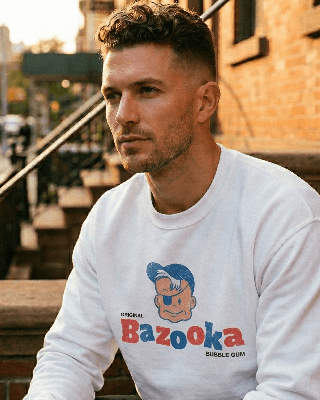 Man wearing BAZOOKA JOE SWEATER | RETRO BUBBLEGUM CLUB in urban setting, showcasing vintage pop culture design.