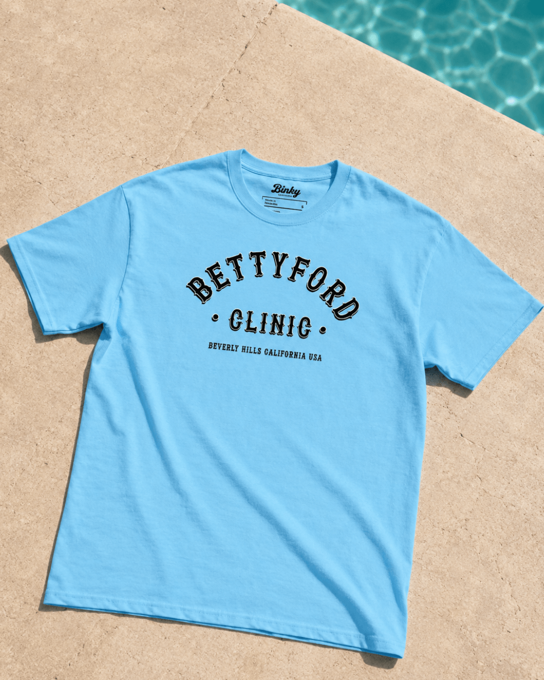 Betty Ford Clinic T-Shirt | Varsity Team in blue, designed by Binky, on a poolside surface.