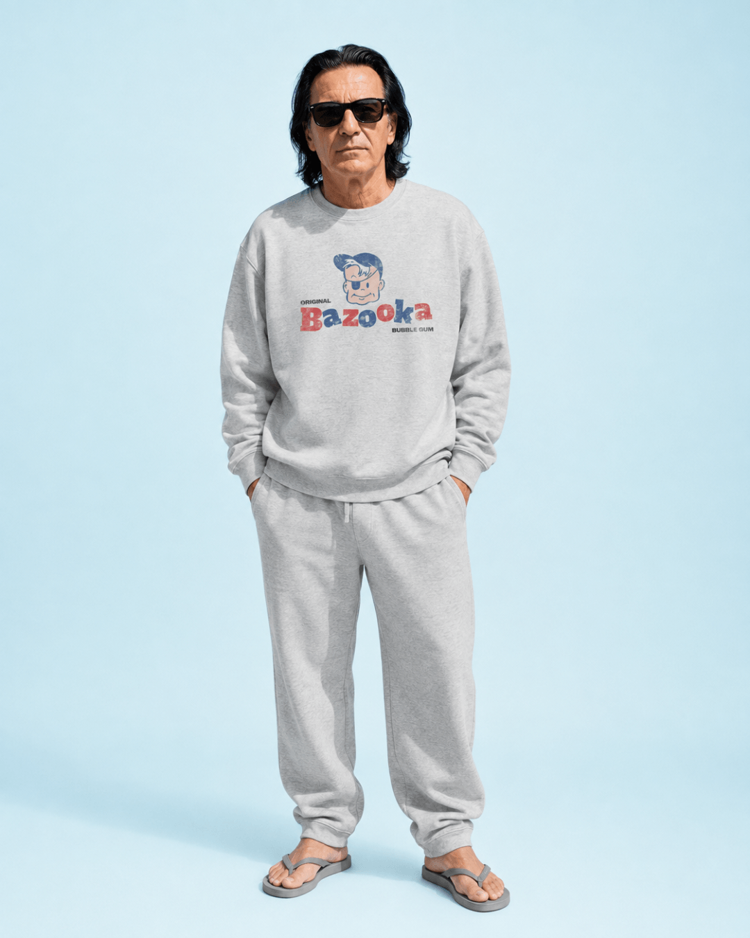 Man wearing a gray Bazooka Joe Club Heavy Sweater with casual pants and sunglasses against a blue background.