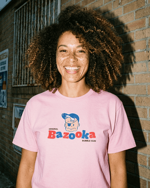 BAZOOKA JOE T-SHIRT | RETRO BUBBLEGUM LEGEND worn by a smiling woman with curly hair in front of a brick wall.
