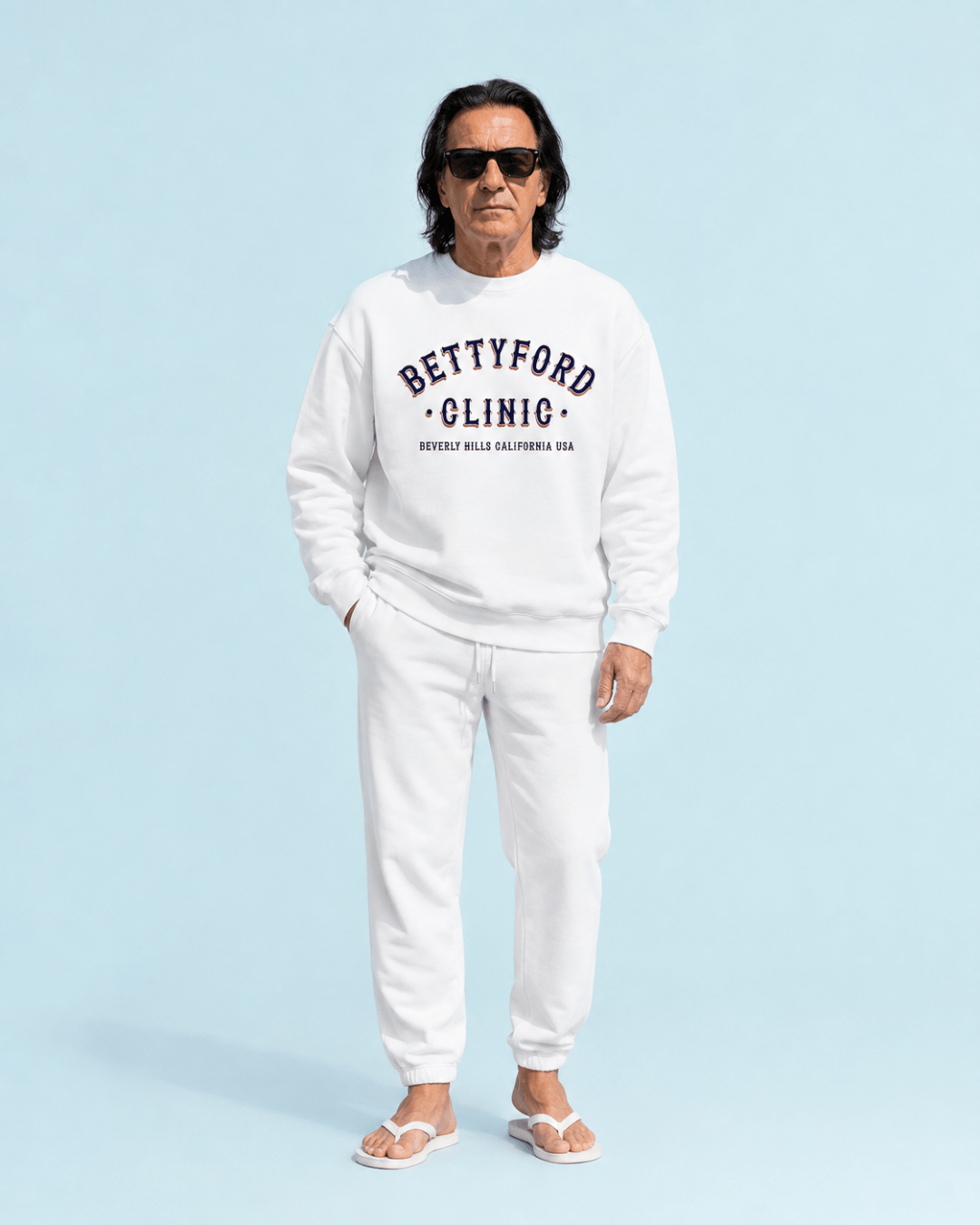 Man wearing a Betty Ford Clinic RWB Heavy Sweater with Beverly Hills print, standing against a blue background.