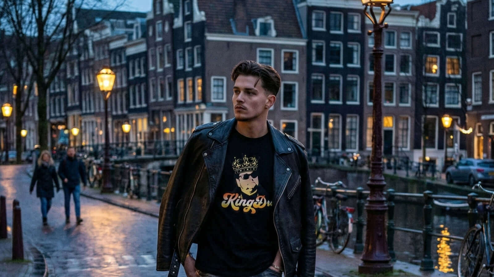 Man in a leather jacket wearing a KING DRE T-SHIRT, walking along a canal in Amsterdam at dusk.