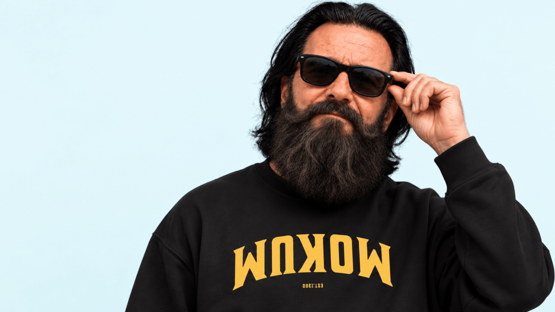 Man with a beard wearing sunglasses and a black sweatshirt, posing confidently against a light blue background.