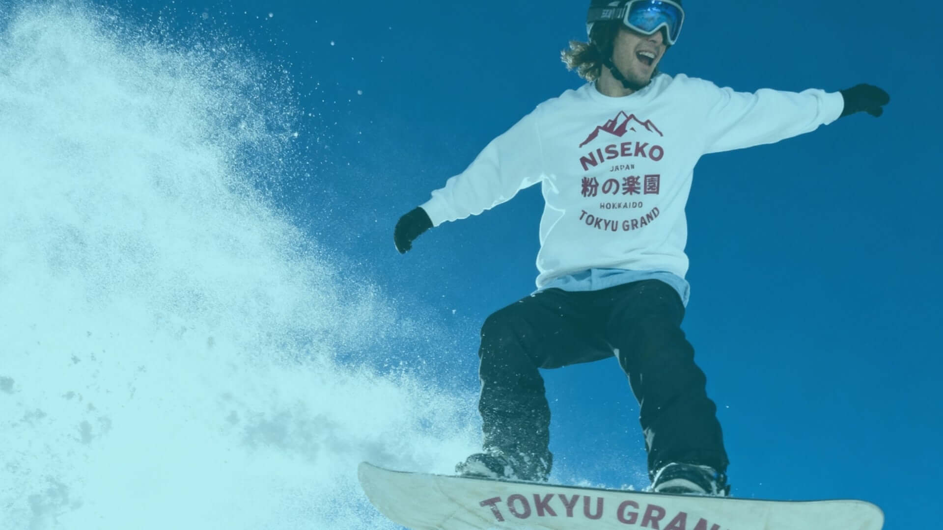 Snowboarder performing tricks in Niseko, wearing Tokyu Grand sweatshirt, surrounded by snow and blue sky.