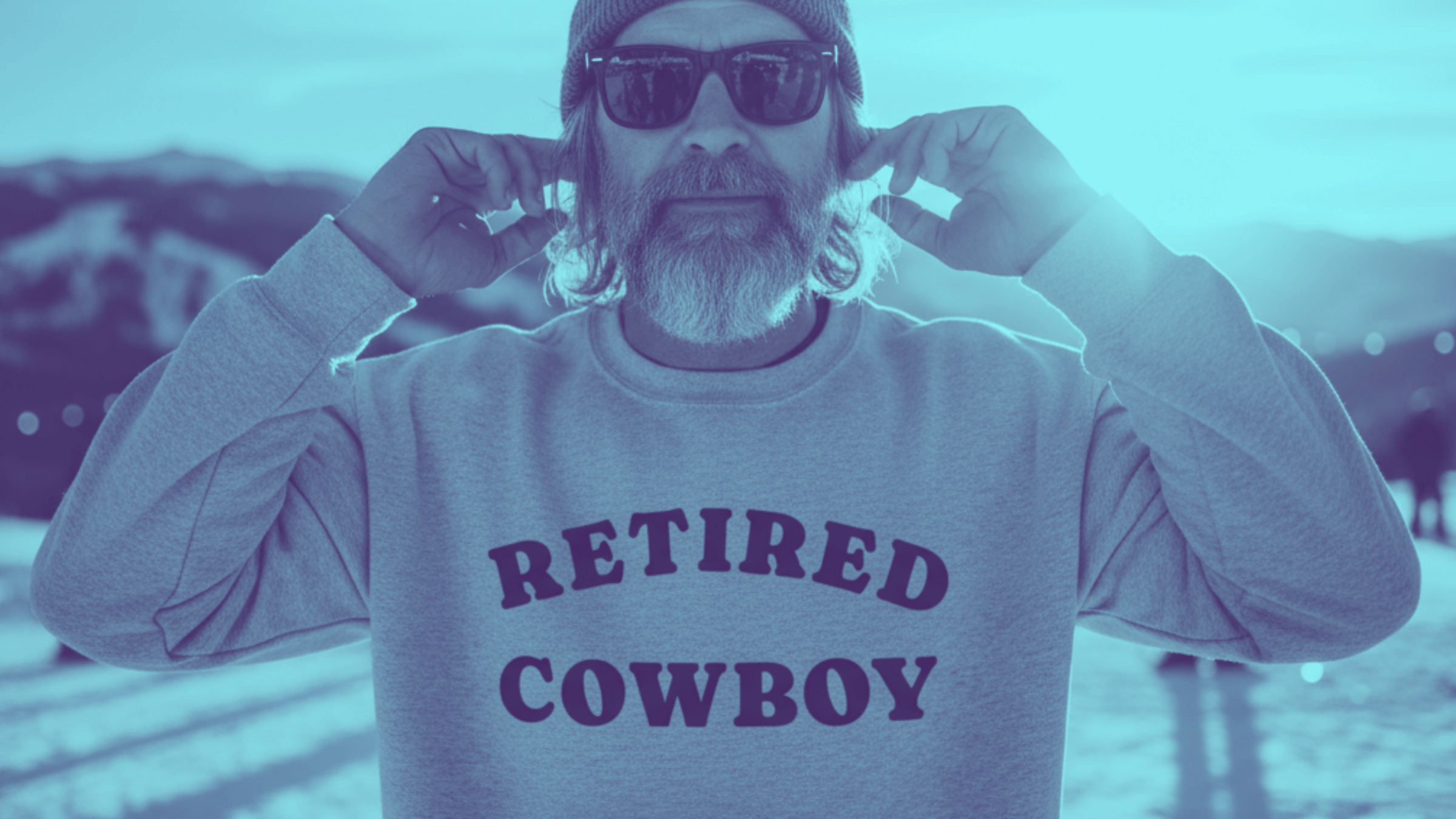 Man wearing a 'Retired Cowboy' sweatshirt in a snowy landscape with mountains in the background.