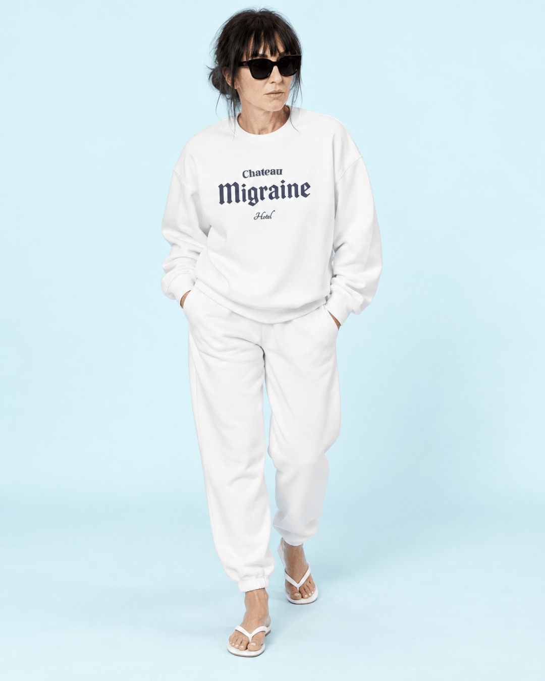 Woman wearing a white Hotel Migraine Heavy Sweater with sunglasses on a light blue background.