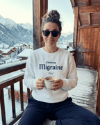 Model wearing HOTEL MIGRAINE HEAVY SWEATER while sitting on a balcony with snow-covered mountains in the background.