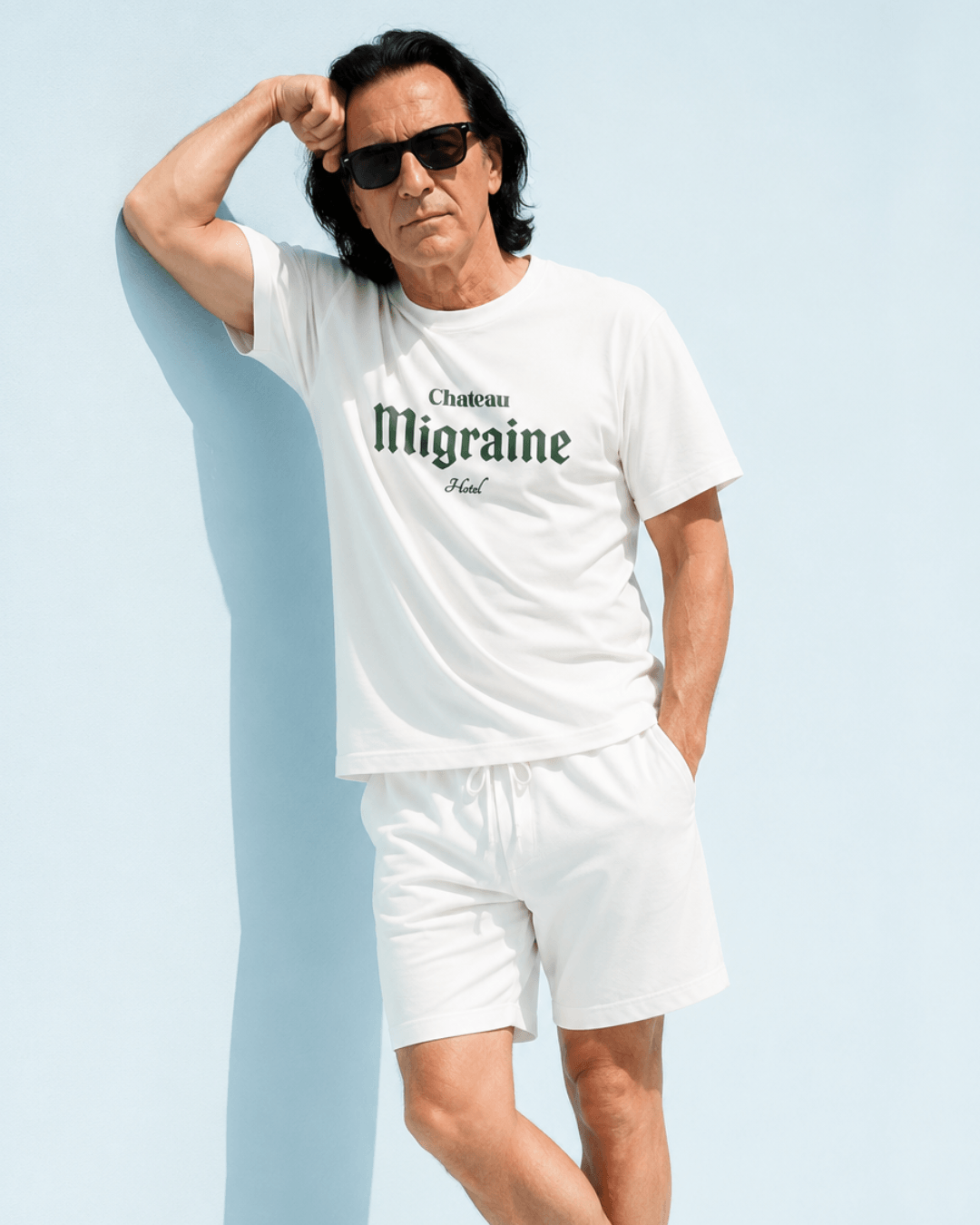 Man wearing Chateau Migraine T-Shirt | Grand Cru Hangover with sunglasses and shorts, against a light blue background.