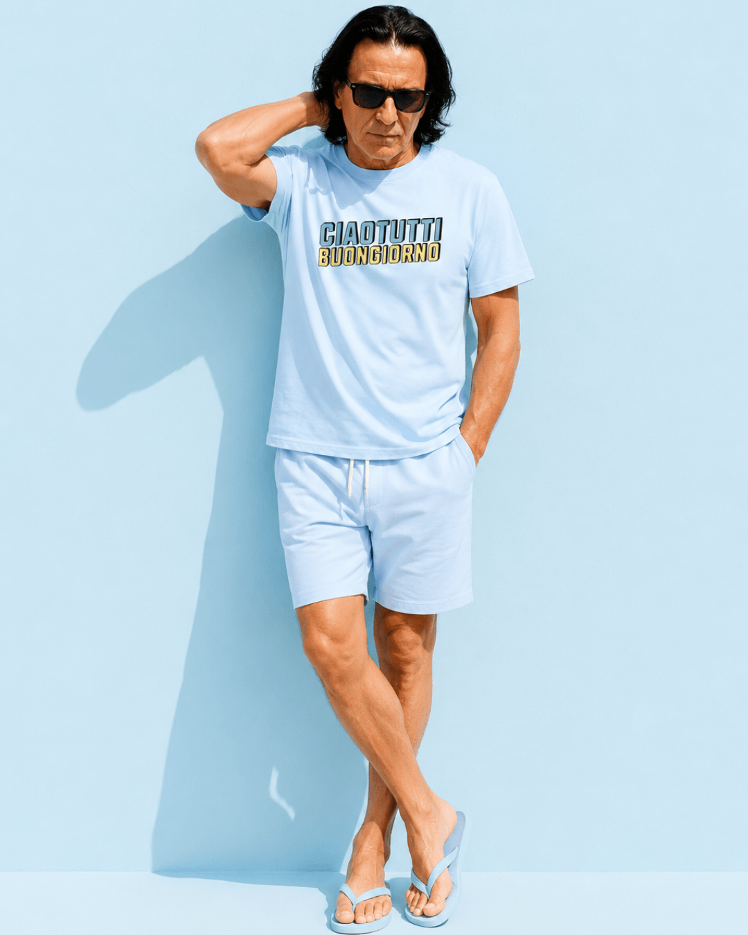 Ciao Tutti T-shirt | Buongiorno Classic worn by man in light blue shorts and flip-flops against a blue background.