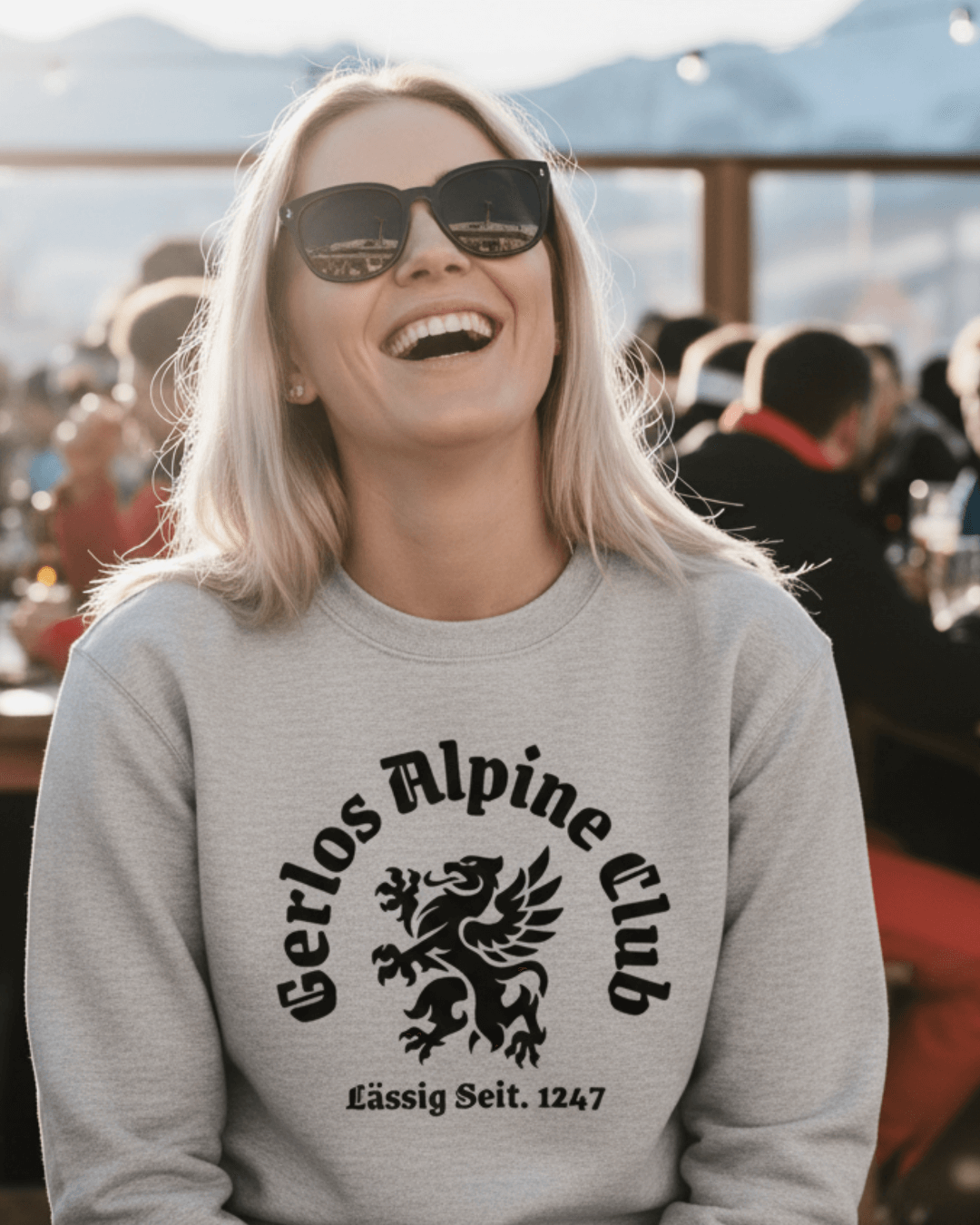 Smiling woman wearing Gerlos Alpine Club Heavy Sweater with griffin crest, enjoying après-ski vibes outdoors.
