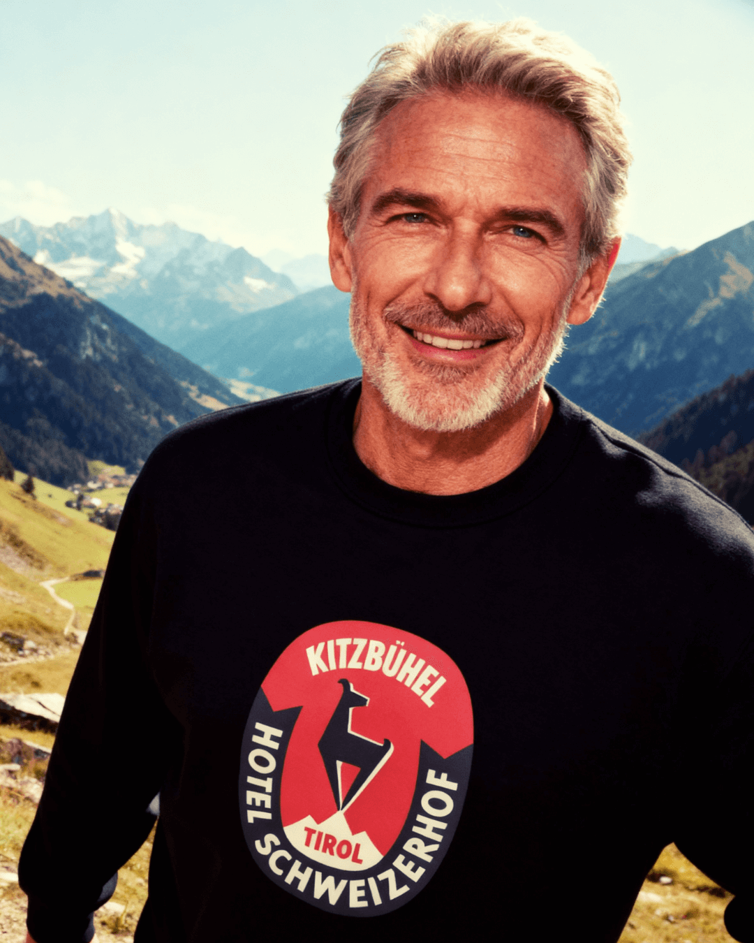 A man wearing the HOTEL KITZBÜHEL HEAVY SWEATER with a retro ski design, set against an alpine backdrop.