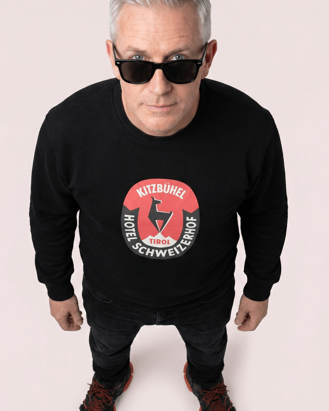 Model wearing HOTEL KITZBÜHEL HEAVY SWEATER with vintage hotel crest against pink background.