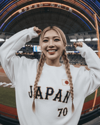 Smiling woman wearing JAPAN 70 BASEBALL SWEATER at a baseball stadium, showcasing vintage style and ultra-soft interior.