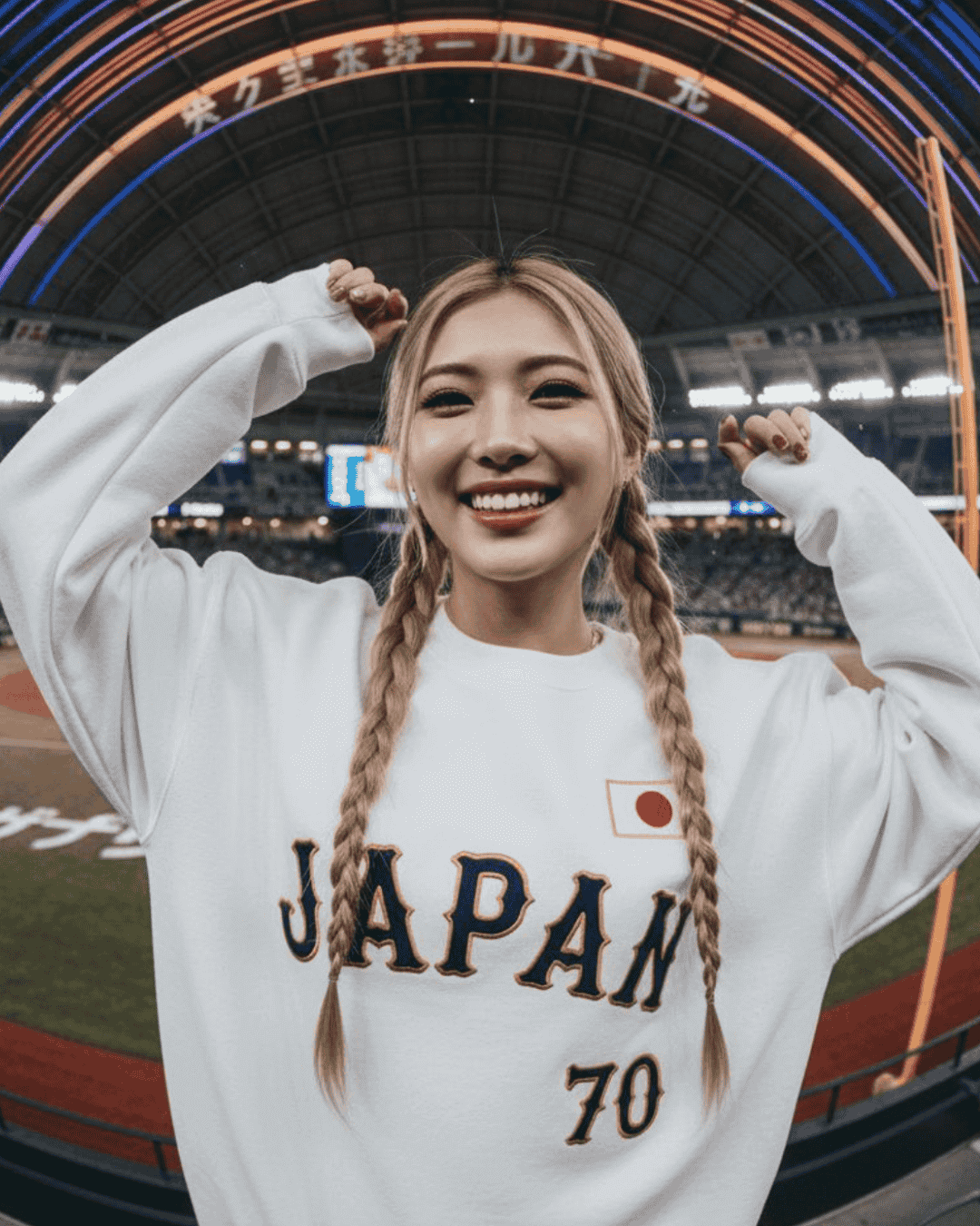 Smiling woman wearing JAPAN 70 BASEBALL SWEATER at a baseball stadium, showcasing vintage style and ultra-soft interior.