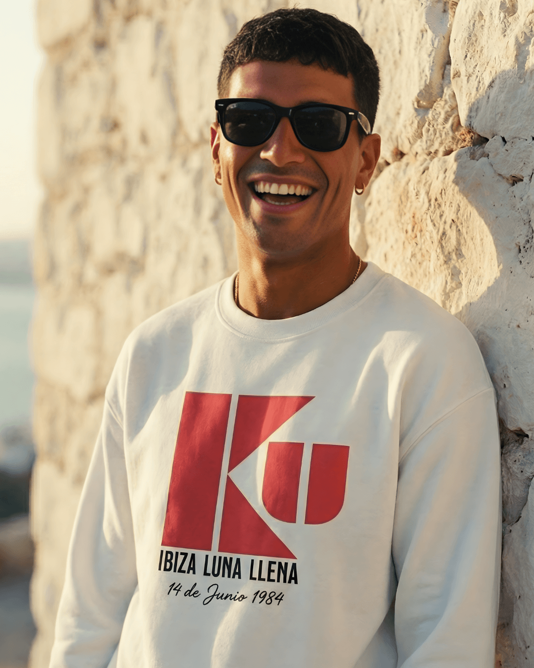 Model wearing KU IBIZA SWEATER | 1984 CLUB LEGEND, smiling outdoors with sunglasses against a stone wall.