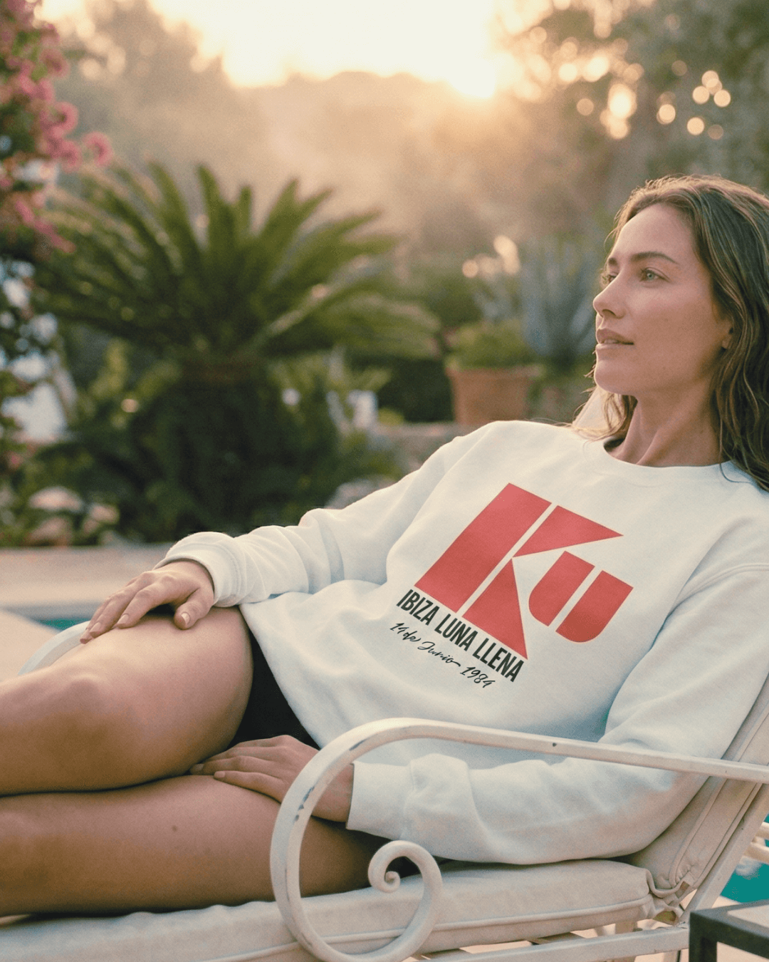 Woman lounging by the pool wearing the KU IBIZA SWEATER | 1984 CLUB LEGEND with iconic logo and sunset backdrop.