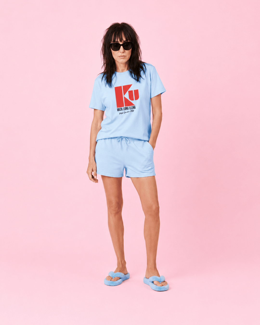 Ku Ibiza T-Shirt | 1984 Club Legend worn by a model in a blue outfit against a pink background.
