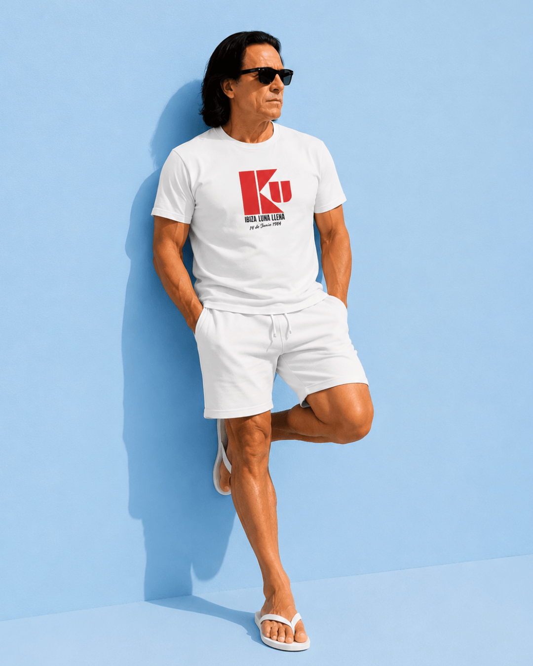 A man wearing a white Ku Ibiza T-Shirt | 1984 Club Legend with sunglasses, leaning against a blue wall.