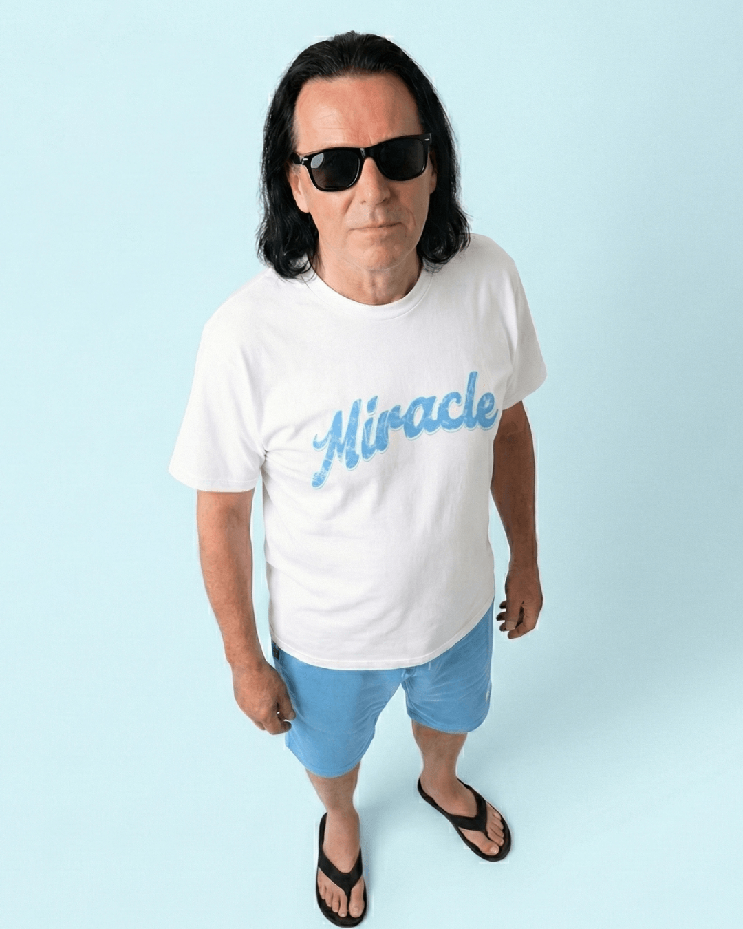 Man in sunglasses wearing a MIRACLE T-SHIRT | LUCK IS EARNED, standing on a light blue background.