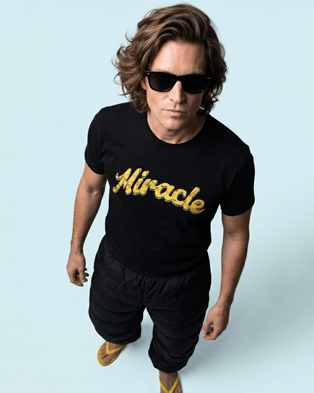 Man wearing MIRACLE T-SHIRT | LUCK IS EARNED, looking cool in sunglasses against a light blue background.
