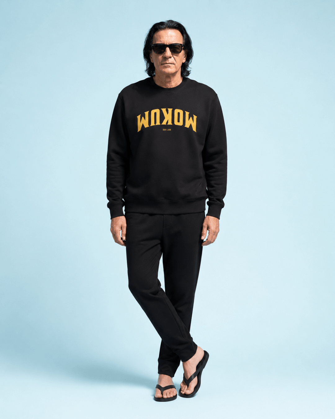 Mokum Est 1300 Heavy Sweater worn by a man in front of a blue background, styled with casual black pants.