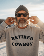 Man wearing a RETIRED COWBOY SWEATER with sunglasses and a beanie, enjoying a sunny outdoor setting.