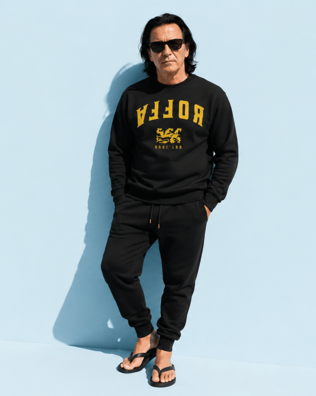 Stylish man wearing Rotterdam Sweater | Roffa 1340 Edition in black with sunglasses standing against a blue background.
