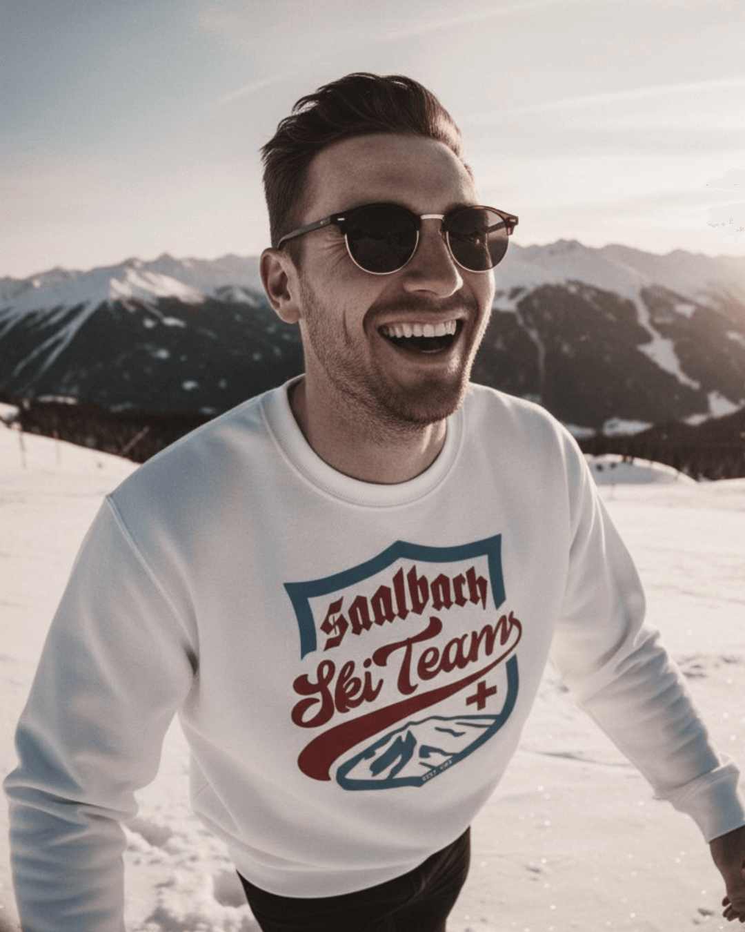 Young man smiling in the mountains wearing a SAALBACH SKI TEAM HEAVY SWEATER, perfect for après-ski and winter vibes.
