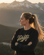 Person wearing a SCHLITZ HEAVY SWEATER against a mountain backdrop during sunset.