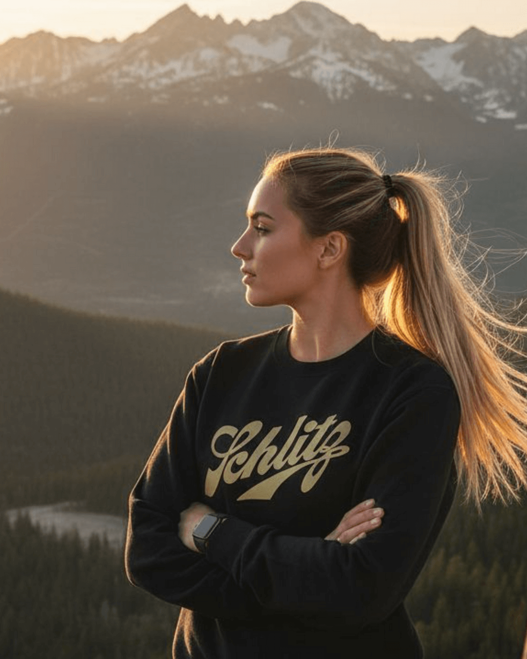 Person wearing a SCHLITZ HEAVY SWEATER against a mountain backdrop during sunset.
