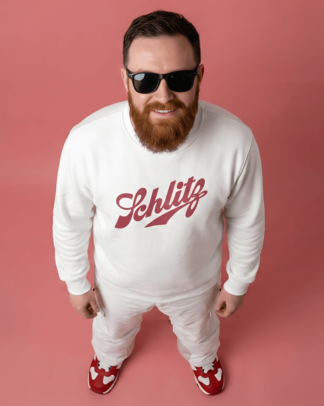 Man wearing SCHLITZ HEAVY SWEATER with sunglasses standing against a pink background.