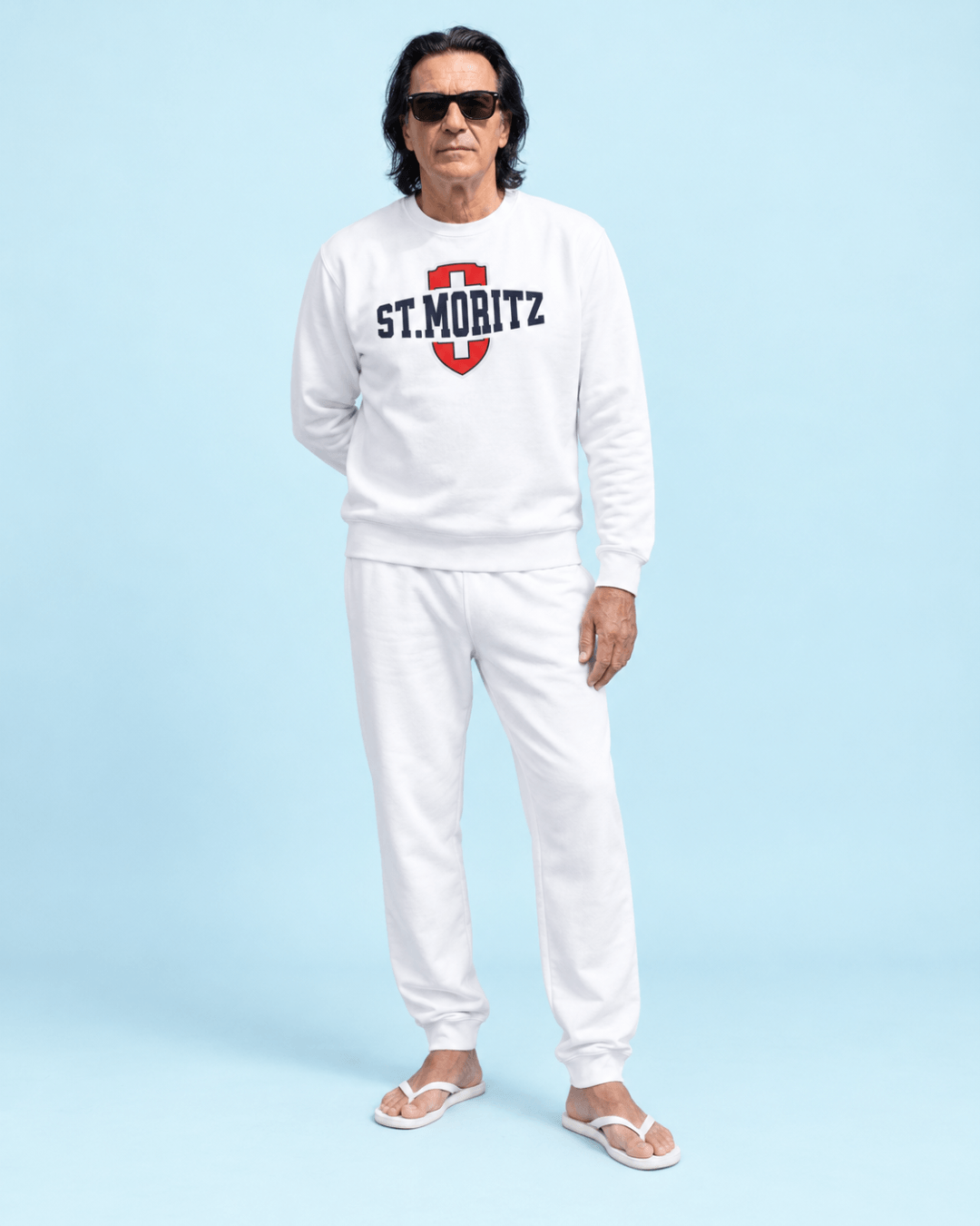Man wearing St. Moritz Heavy Sweater and white pants, showcasing Swiss style on a light blue background.