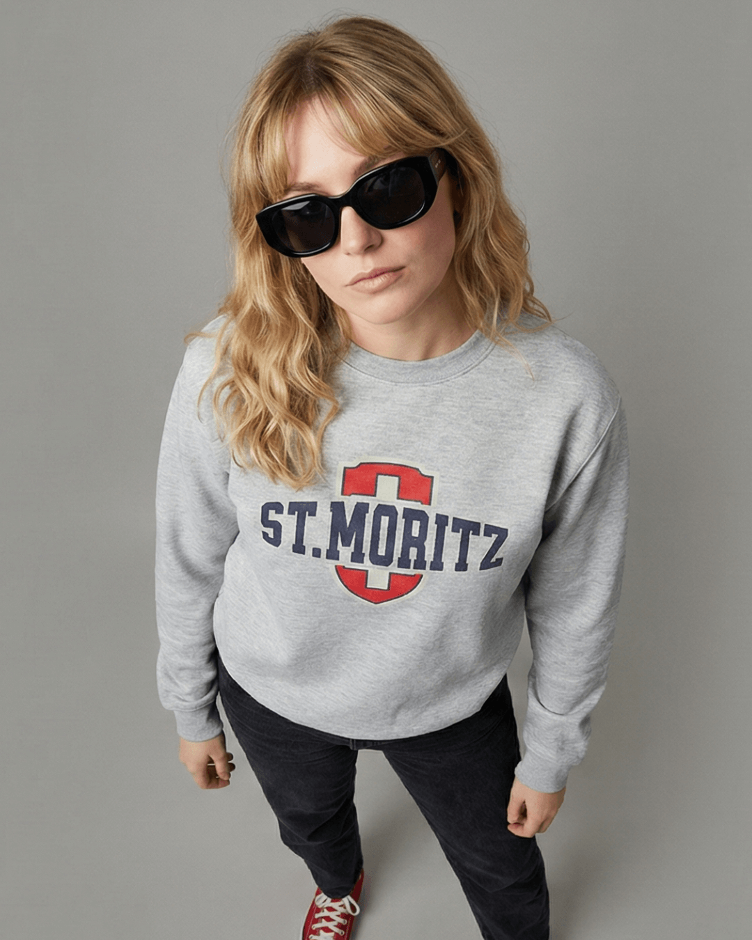 A model wearing the ST. MORITZ HEAVY SWEATER in grey with a bold logo and stylish sunglasses.