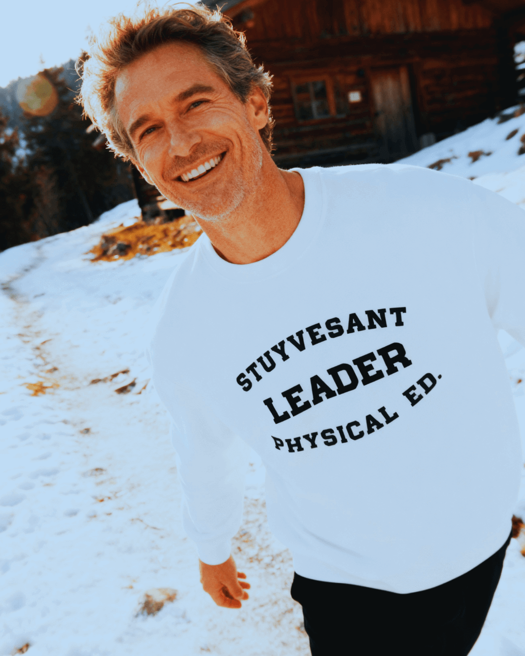 A smiling man wearing a STUYVESANT LEADER HEAVY SWEATER with the text 'STUYVESANT LEADER PHYSICAL ED.' in a snowy landscape.