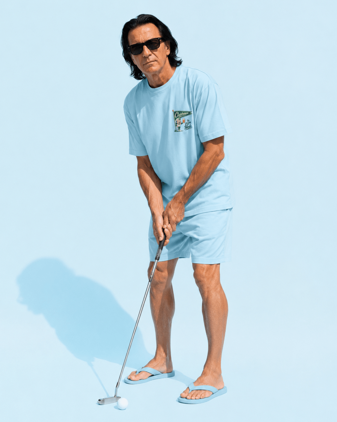 A man wearing The 19th Hole T-Shirt | Clubhouse Classic with sunglasses, putting golf ball on a blue background.