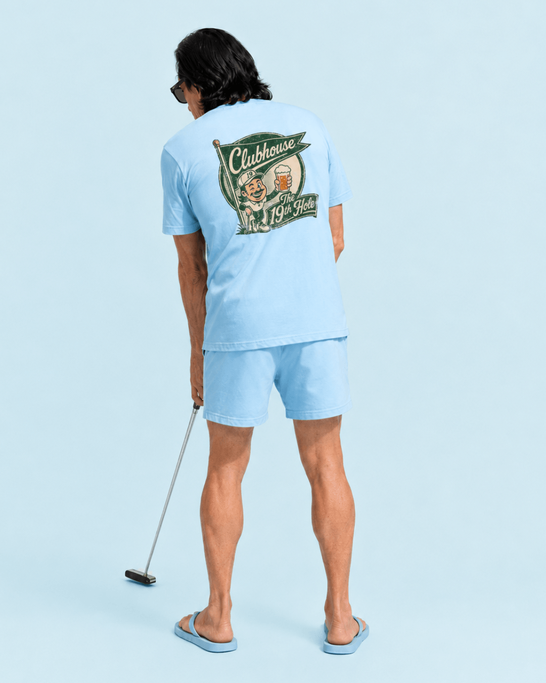 A man wearing The 19th Hole T-Shirt | Clubhouse Classic, playing golf with a putter, showcasing the design on the back.