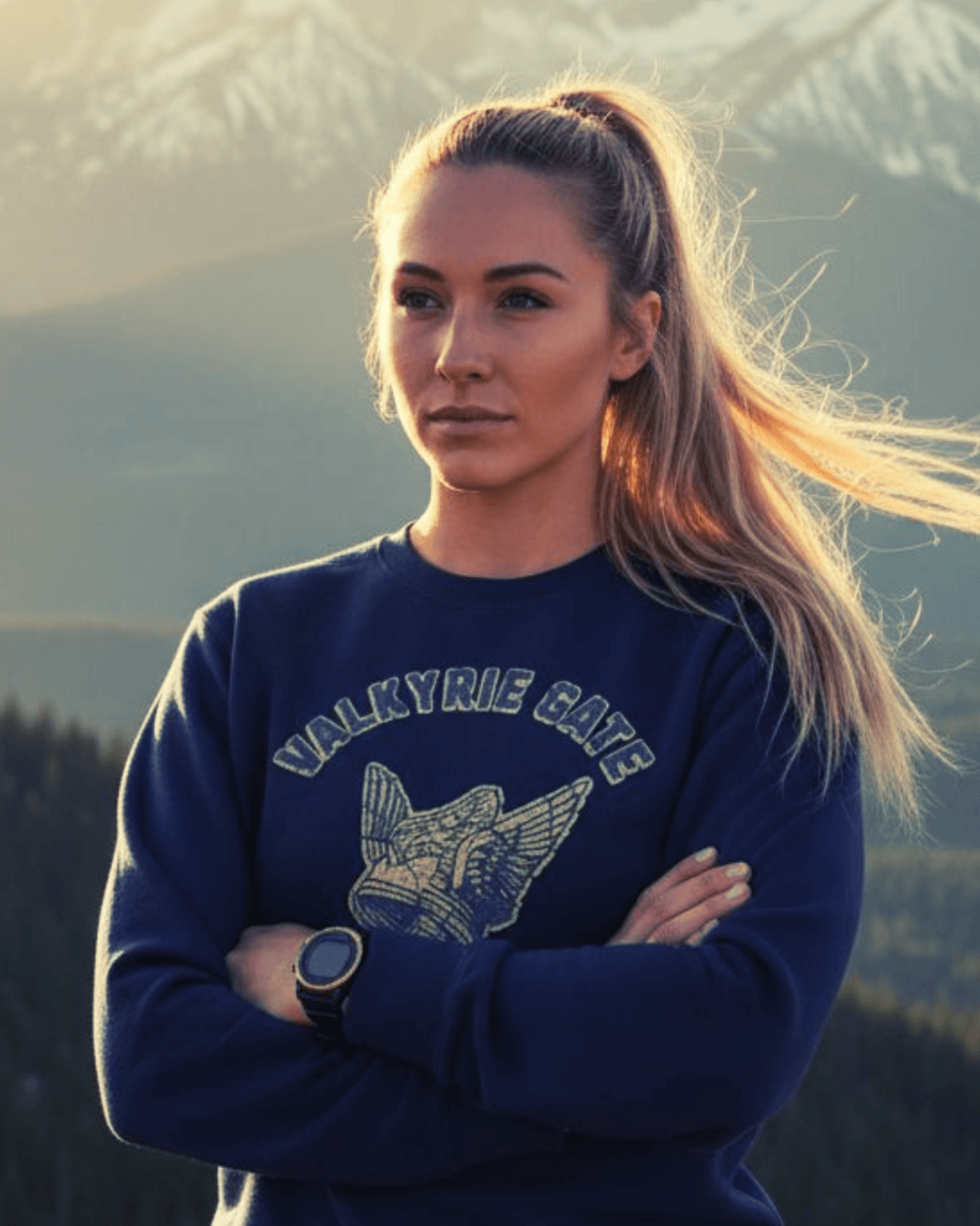 Young woman wearing VALKYRIE GATE HEAVY SWEATER with a winged helmet emblem in a mountainous outdoor setting.